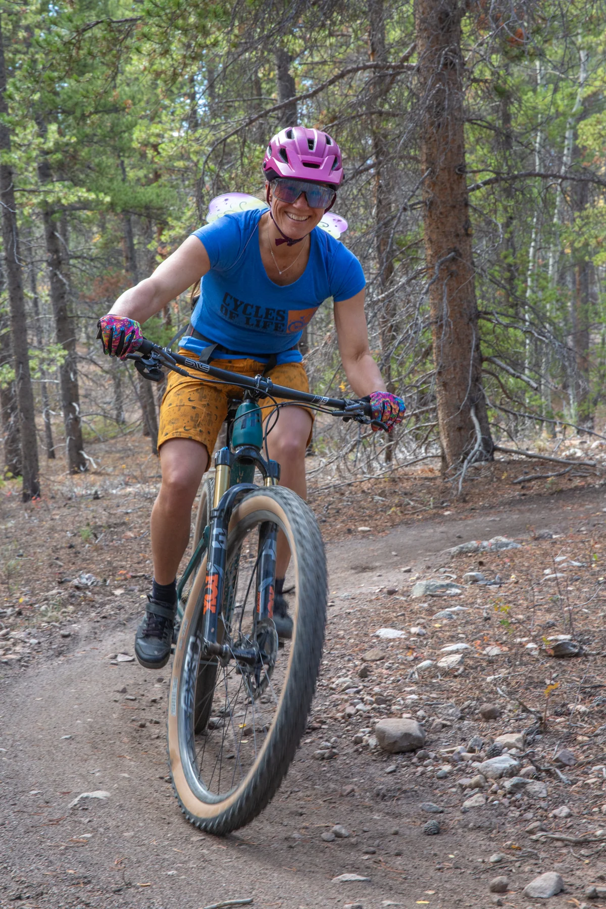 A smiling cyclist in a pink helmet and blue shirt rides through the forest wearing fairy wings.