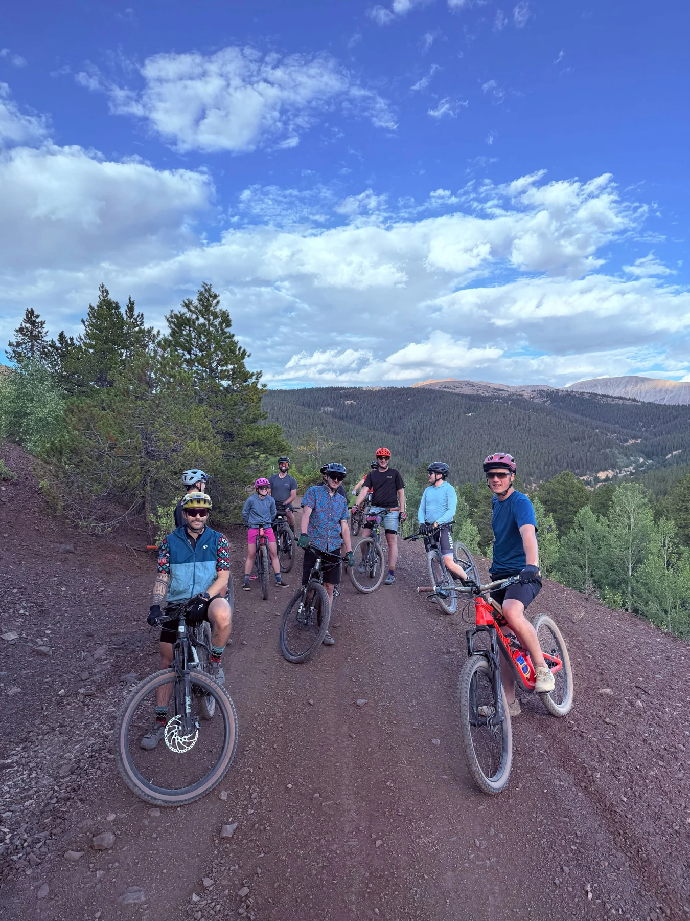 Cyclists gather with their bikes on a mountain trail under a blue, cloud-filled sky.