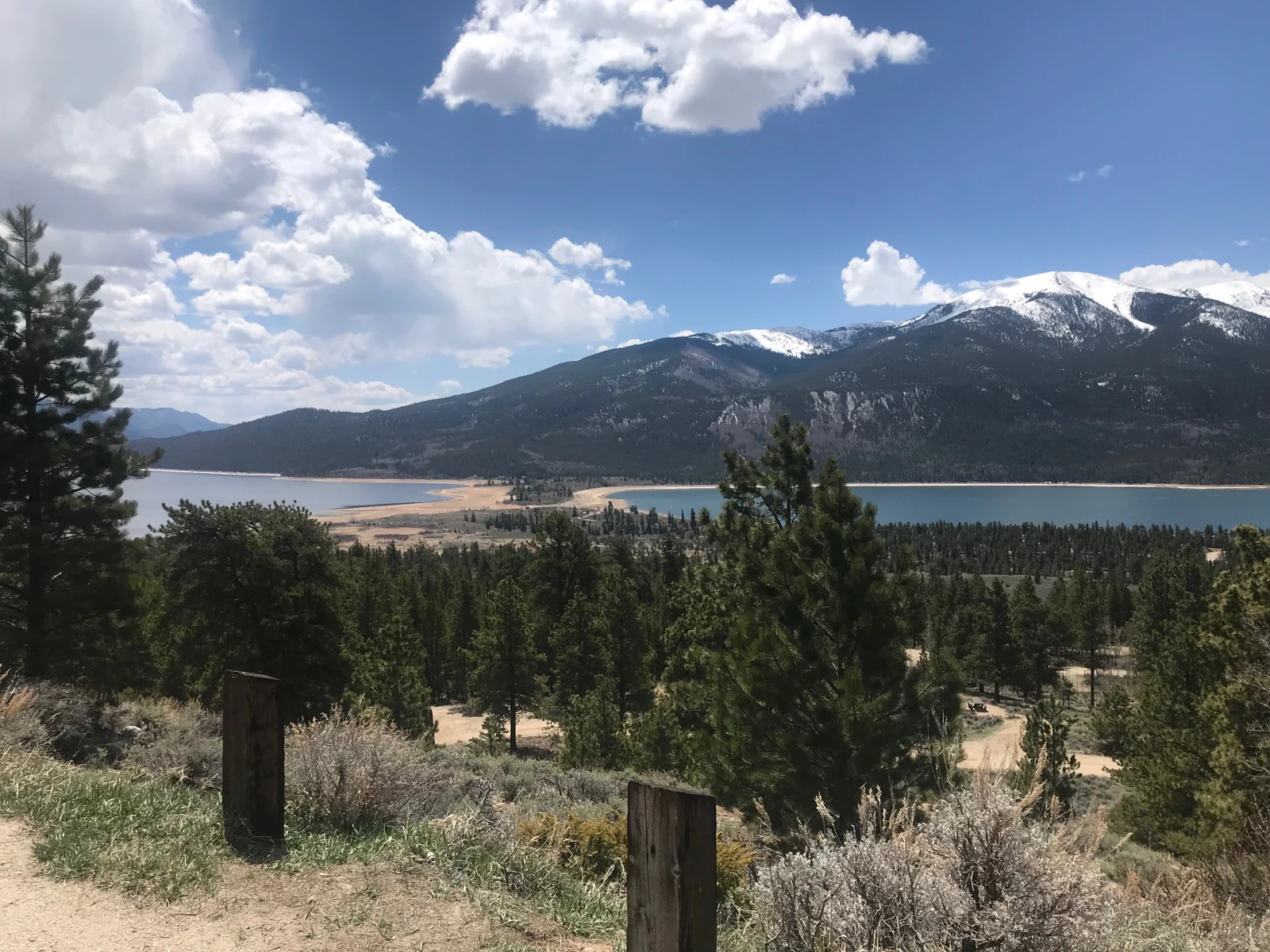 Twin Lakes and a snow-covered mountain ridge appear beyond pine trees beneath a cloud-filled spring sky.