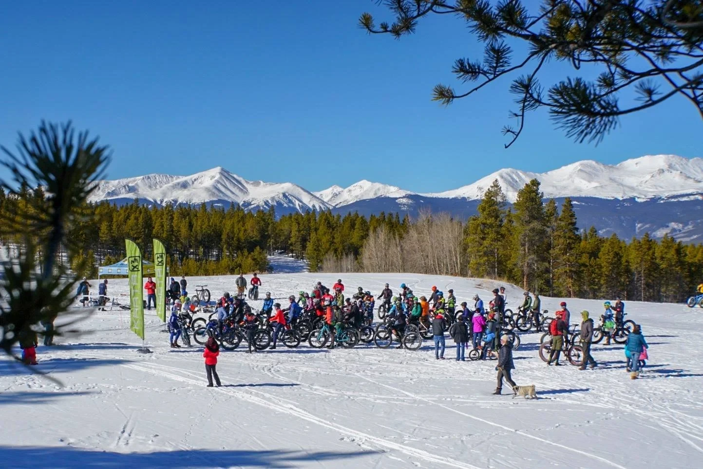 Fatty Patty riders assemble at a sunny snowy start area below mountain peaks.