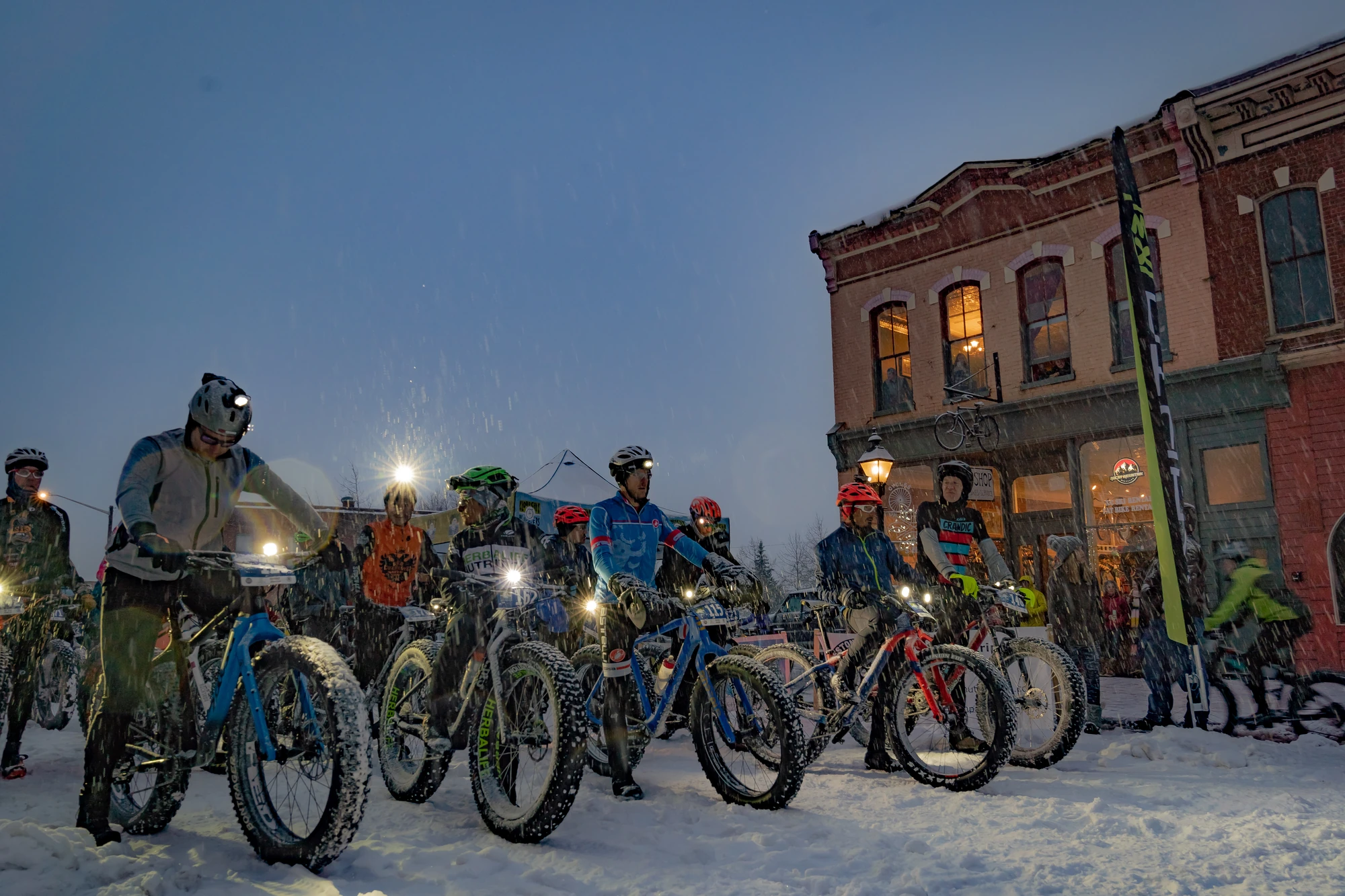 Mineral Belt Mayhem racers line up on snowy Harrison Avenue at dusk beside the Cycles of Life storefront.