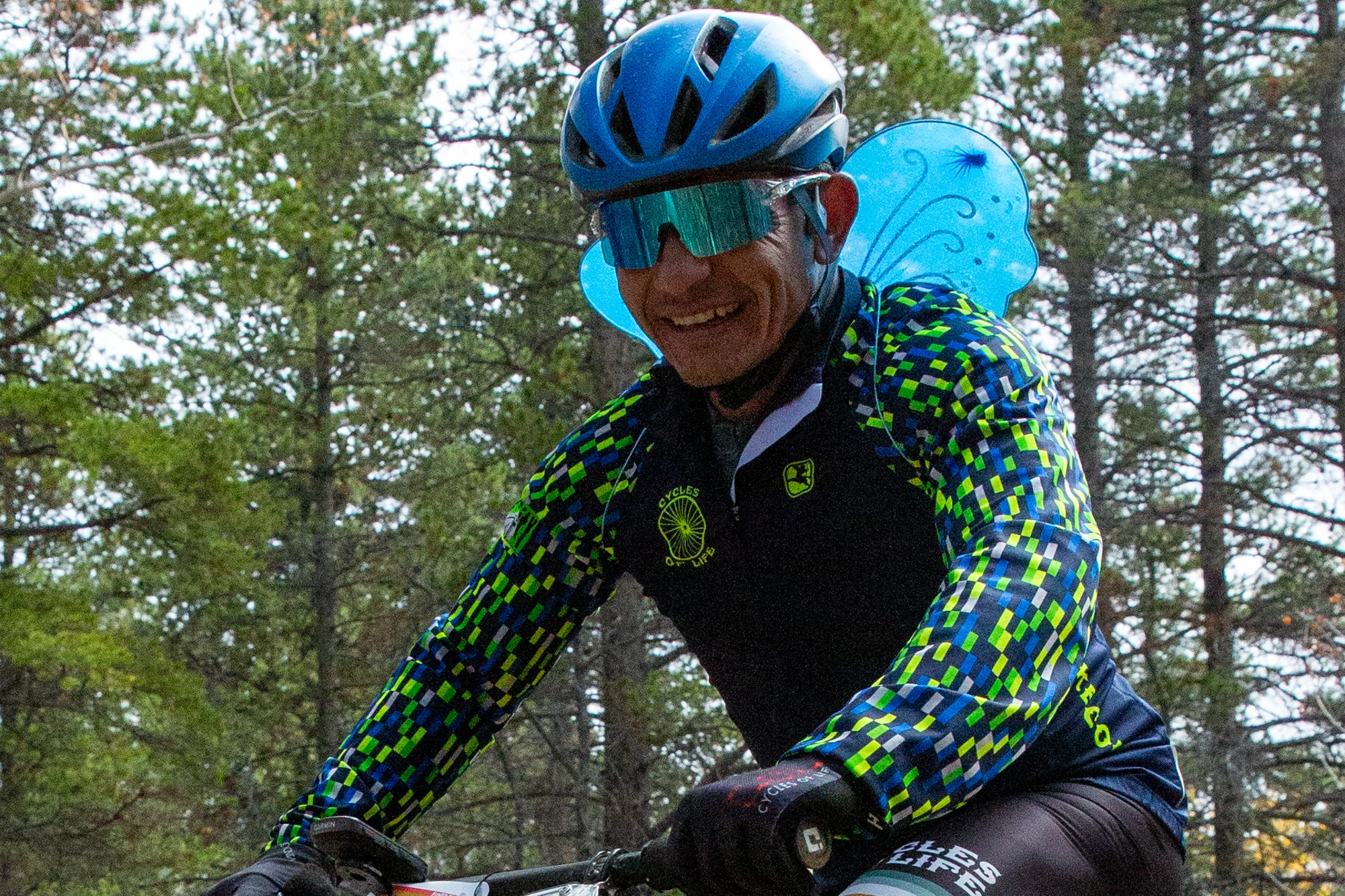 Smiling rider with blue helmet and fairy wings on a wooded trail.