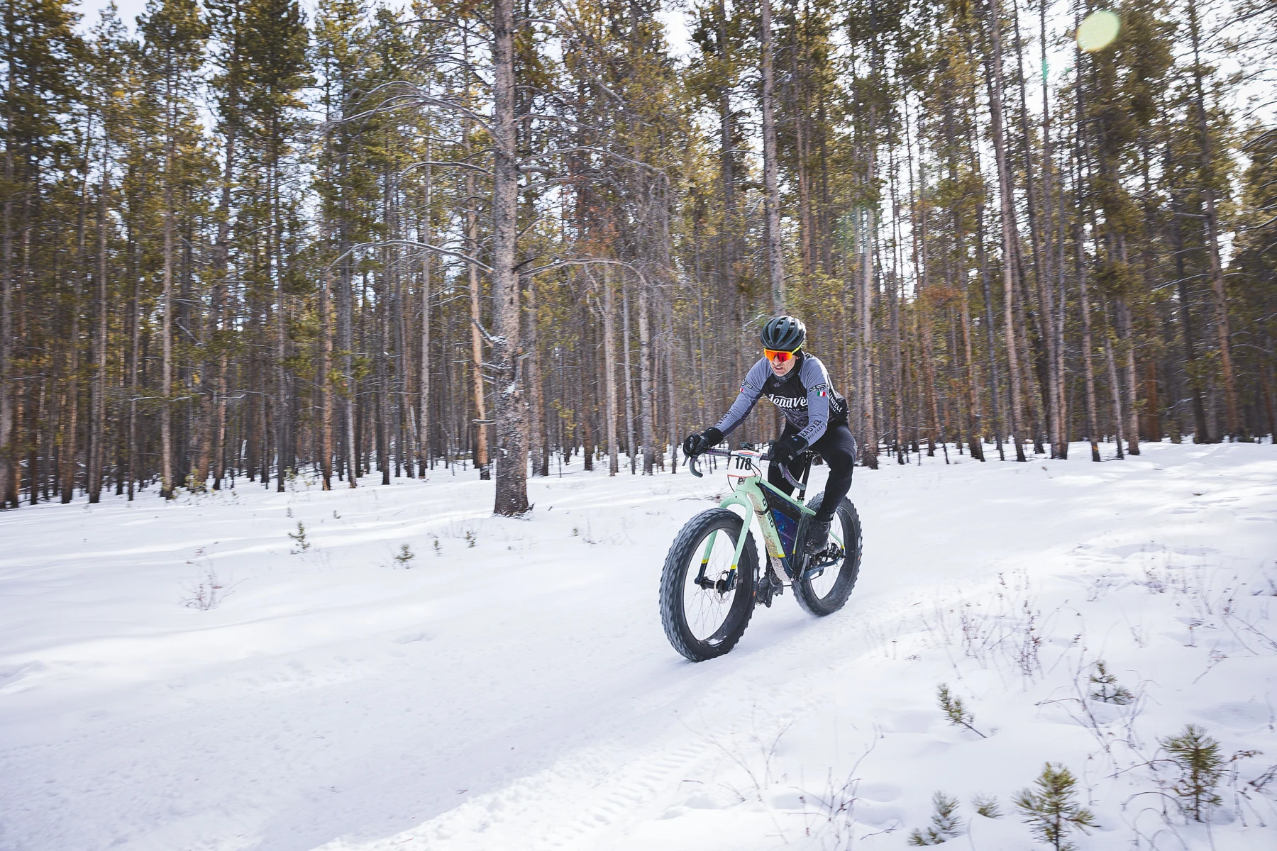 A fat-bike rider rounds a snowy forest trail in bright winter sunshine.